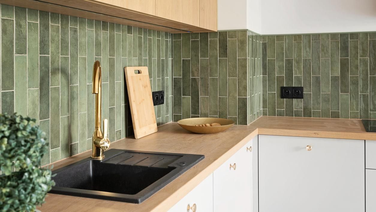 Et moderne kjøkken med grønne fliser, sort vask, messingkran og benkeplate i treimitasjon.