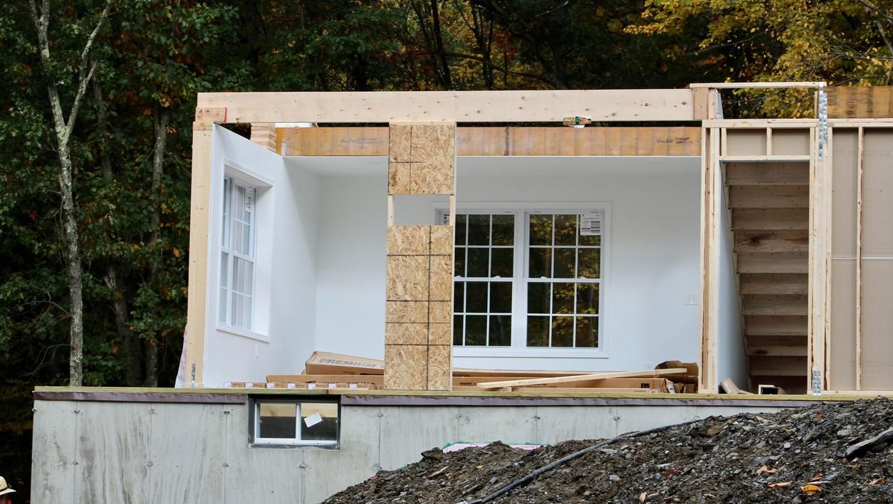 Et hus under bygging med synlig trekonstruksjon, åpne vegger og vinduer montert i en hvitmalt del.